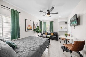 This inviting studio combines comfort and style, featuring a queen-size bed, a cozy leather sofa, and a dining area for two. The space is enhanced by vibrant wall art, natural light from windows, and soft greenery, creating a relaxing atmosphere.
