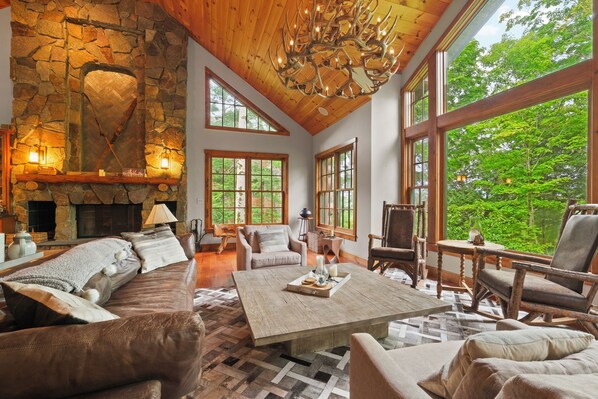 Bright and inviting living space with a stunning stone fireplace, cozy seating, and large windows offering breathtaking forest views. The perfect spot to relax and unwind in nature, complete with rustic decor and a grand chandelier.