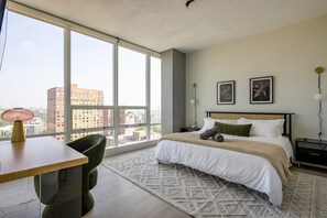 Modern bedroom with floor-to-ceiling windows with a city view. It features a bed with white linens, green and beige accents, a wooden desk, a green chair, and two floral prints above the bed. Rug, full length mirror and 55" TV and Private Bathroom
