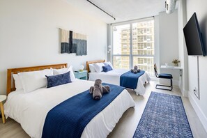 Bright bedroom with two queen beds featuring white linens and blue accents. A wall tapestry decorates the space above the beds. Large windows offer a city view, and a wall-mounted 55" TV, full length mirror and folding desk with a chair