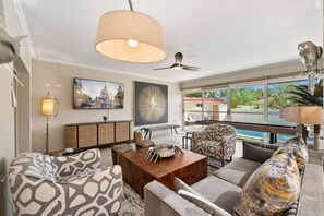 - Modern living room with patterned armchairs and a wooden coffee table - Glass doors fill the space with natural light, offering a clear view of the outdoor pool area - Stylish decor includes abstract art, a mounted TV, and elegant lighting fixtures