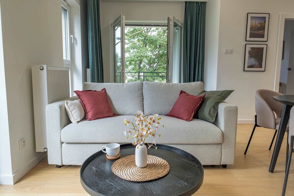 A stylish living room with a light sofa and soft cushions invites you to relax with a coffee, while the large window fills the space with natural light.