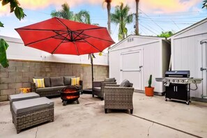 outdoor patio with gray wicker furniture, a red umbrella, a fire pit, white storage sheds and a barbeque grill