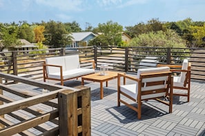 Outdoor patio area featuring modern wooden furniture with white cushions. The scene is calm and inviting, with green trees visible in the background.