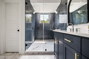 A glass-enclosed shower with a dark, vertically stacked subway tile wall and a rainfall showerhead. The flooring is a white marble pattern, adding a touch of elegance to the overall design.