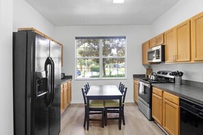 The kitchen features cabinets, a four-burner stove with an oven, a microwave, a kettle, a large refrigerator, and a dishwasher. It also includes a dining table for four, a bar area, and natural light that fills the space, creating a bright and welcoming atmosphere.