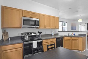 The kitchen features cabinets, a four-burner stove with an oven, a microwave, a kettle, a large refrigerator, and a dishwasher. It also includes a dining table for four, a bar area, and natural light that fills the space, creating a bright and welcoming atmosphere.