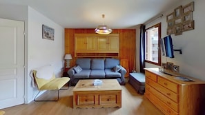 The living area is furnished with a blue sofa, a yellow armchair, a coffee table, a wooden dresser, a flat screen TV, and a window overlooking the ski resort.This is a cozy and comfortable space for relaxing after a day of skiing.