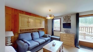 This inviting living area features a comfortable sofa, a coffee table, and a flat-screen TV. Sliding doors lead to a private terrace with a view of the surrounding mountain landscape. The warm wood paneling on the walls adds a touch of rustic charm to th