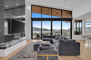 This modern living room boasts floor-to-ceiling windows with panoramic lake and mountain views. A dark gray sectional, marble fireplace, and sleek dining area create a luxurious, airy space with abundant light.
