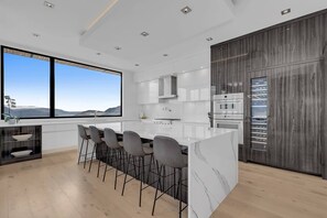 This modern kitchen features a marble island with six grey bar stools, stainless steel appliances, and dark wood cabinetry. A large window offers panoramic lake and mountain views, with recessed lighting and light hardwood floors.