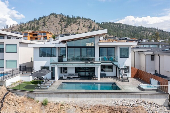 This modern hillside retreat features sleek white architecture, large windows, and breathtaking lake and mountain views. Enjoy outdoor seating, an infinity pool, and hot tub surrounded by lush greenery.
