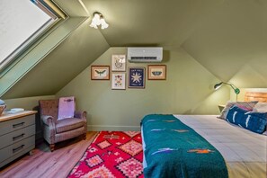 Queen bed in the upstairs bedroom with AC, black out blinds, storage & a cozy vintage chair.