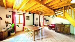 The open-plan living area features a charming dining table with four chairs, ideal for enjoying meals together. Natural light streams through large windows, illuminating the space and highlighting the rustic wooden beams that adorn the ceiling. A comforta