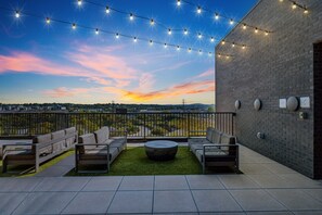 Rooftop patio with string lights, modern outdoor furniture, and a central fire pit, overlooking a scenic sunset view with a mix of urban and natural landscapes.