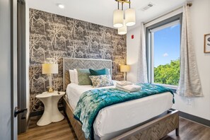 A modern bedroom with a double bed, patterned wallpaper, two bedside tables with lamps, and a large window with a view of greenery. The bed has folded towels on top.