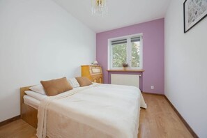 A cozy bedroom with white walls, a light wood bedframe, and beige bed linens. The room feels airy with plenty of natural light.