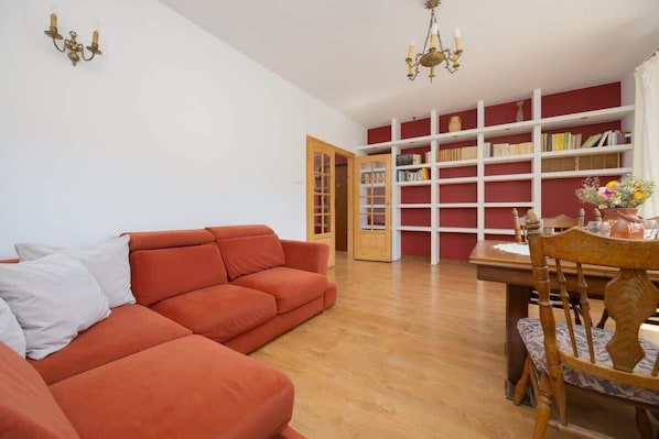 A spacious living room with light wooden floors, red armchairs, and table. The large bookshelf in the background adds warmth to the room.