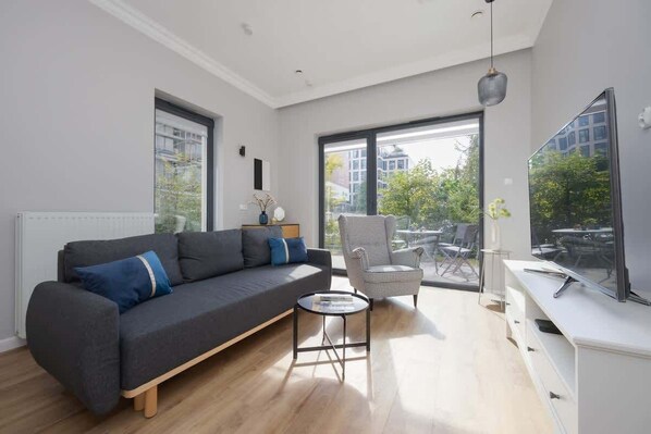 A bright living room with a dark gray sofa, a small coffee table, and large floor-to-ceiling windows, offering a beautiful view of the outdoor greenery and ample natural light.