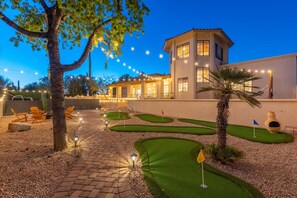 As twilight sets in, your backyard transforms into a golfer’s paradise. The mini-golf course, illuminated by string lights and ground lights, offers a fun and relaxing escape. The cozy ambiance, with the two-story house in the background and the beautifully lit trees, makes it a perfect spot for evening leisure. Pure bliss!
