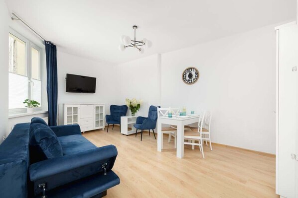 A cozy living room with blue armchairs, a white table, and a TV mounted on the wall. The space is bright, with wooden floors and minimalistic decor.