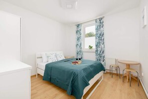 A bedroom featuring a bed with teal bedding and floral curtains. The room has a small wooden desk by the window.
