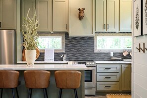 Modern cabin kitchen with sage green cabinets, herringbone backsplash, marble island, leather barstools, and stainless steel appliances. Stylish and functional culinary space in a cozy forest retreat.