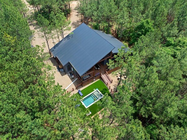 Drone view of luxury cabin surrounded by pine forest, showcasing private pool, patio deck, and nature escape.
