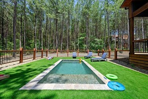 Backyard plunge pool with lounge chairs, cornhole game, and lush artificial turf in a serene forest setting