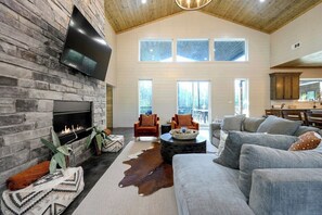 Spacious living room with vaulted ceiling, stone fireplace, comfy seating, and natural light from tall windows.