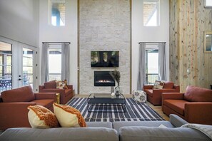 Spacious living room with high ceilings, stone fireplace, modern furnishings, and large windows offering forest views.