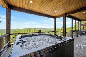 Covered deck with large bubbling hot tub and stunning panoramic views of pine-covered hills.