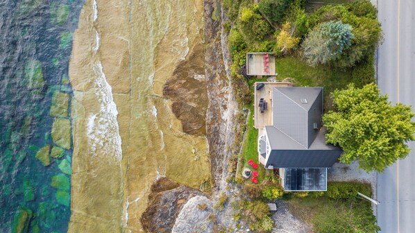 Sure! Here's a more concise version:

---

Aerial view of a modern waterfront home with a spacious deck, fire pit area, and lush greenery, set along a rocky shoreline with clear, shallow waters.