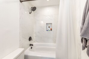 A modern bathroom featuring a white bathtub with a mounted black showerhead and faucet, white marble tiles, a built-in shelf with toiletries, and a light gray towel hanging on the right side.