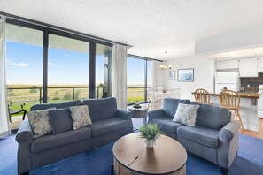 Modern open-concept living space with large windows, blue sofas, a cozy dining area, and a bright, inviting atmosphere. Perfect for relaxation and entertaining.
