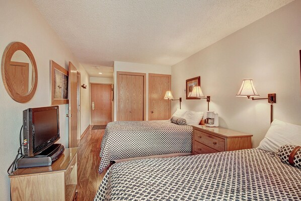 A hotel room with two double beds, a small flat-screen TV on a dresser, wall-mounted lamps, and wooden furnishings. The room has white walls, patterned bed covers, and a wooden floor.