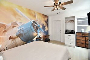 Sleeping area showing microwave oven, ceiling fan, dresser looking at a modern airplane theme painting on wall
