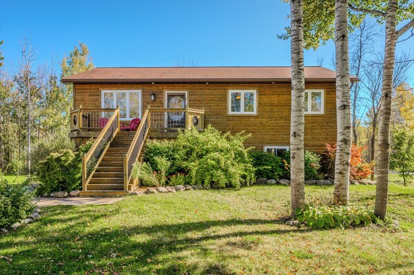 Welcome to Black Bear Cottage, a peaceful outdoor setting with tall trees and soft sunlight. A modern wooden staircase leads to a home with natural wood siding all throughout. 