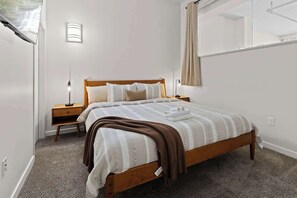 The room features a plush queen bed dressed in soft linens and complemented by warm, neutral tones that create a calming atmosphere. With bedside tables and modern lamps, the space offers both functionality and elegance. 