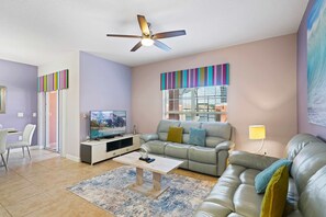 Cozy living room featuring plush leather sofas in soft gray tones, accented with colorful pillows - Striped window valances add a playful touch, complementing the light purple walls - Ceiling fan and modern decor create a welcoming atmosphere