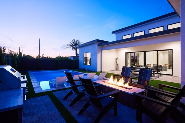 Resort-like backyard with a covered patio offering outdoor dining with seating for 8 guests, a sparkling blue heated pool with sun loungers, BBQ grill and a fire pit.