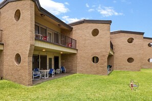 Exterior view of the brick building with private patios on the ground level and balconies on the upper floor, surrounded by a large grassy courtyard.