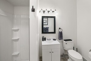 Bathroom features a corner shower with built-in shelves, a white vanity topped by a black-framed mirror and overhead lighting, adjacent wall-mounted towel ring, and a sleek toilet completing the layout. 🚿🪞🧽