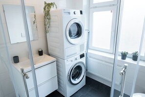 Bright and functional laundry area featuring a stacked washer and dryer, a modern white vanity with a sink, and a wall-mounted mirror. The space is accented with sleek black floor tiles and a touch of greenery, creating a clean and organized environment for everyday tasks.