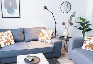 Stylish living room featuring two cozy gray sofas with warm-toned throws and patterned cushions. A sleek white coffee table, floor lamp, and decorative elements like potted plants and a modern wall mirror create an inviting atmosphere. Large windows allow natural light to enhance the bright and contemporary design.