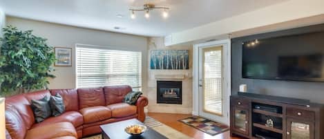 A living room with a red sectional sofa, a coffee table, a wall-mounted TV, a fireplace, a large plant, and windows with blinds. There is also a door leading outside and a rug on the hardwood floor.