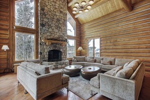 Spacious living room featuring a stone fireplace, large sectional sofas, wooden ceiling with beams, and floor-to-ceiling windows with views of the trees outside.