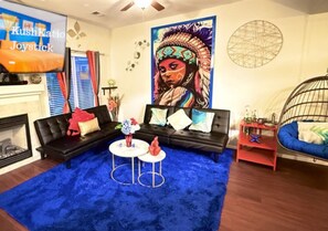 The living room is colorful with a striking mural, black sofas, a blue rug, and a fireplace. A hanging chair and red curtains add to the decor, while a small table with vases and a metal wall