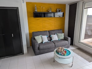 Helles und modernes Wohnzimmer mit einem Schlafsofa für zwei Personen und einem TV. Dank der raumhohen Fenster bietet es einen freien Blick auf die Terrasse mit Blick auf den Yachthafen