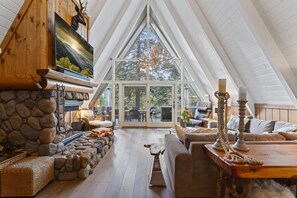 Cozy A-frame cabin interior featuring a stone fireplace, warm wooden accents, and a spacious seating area with a view of the forest beyond.